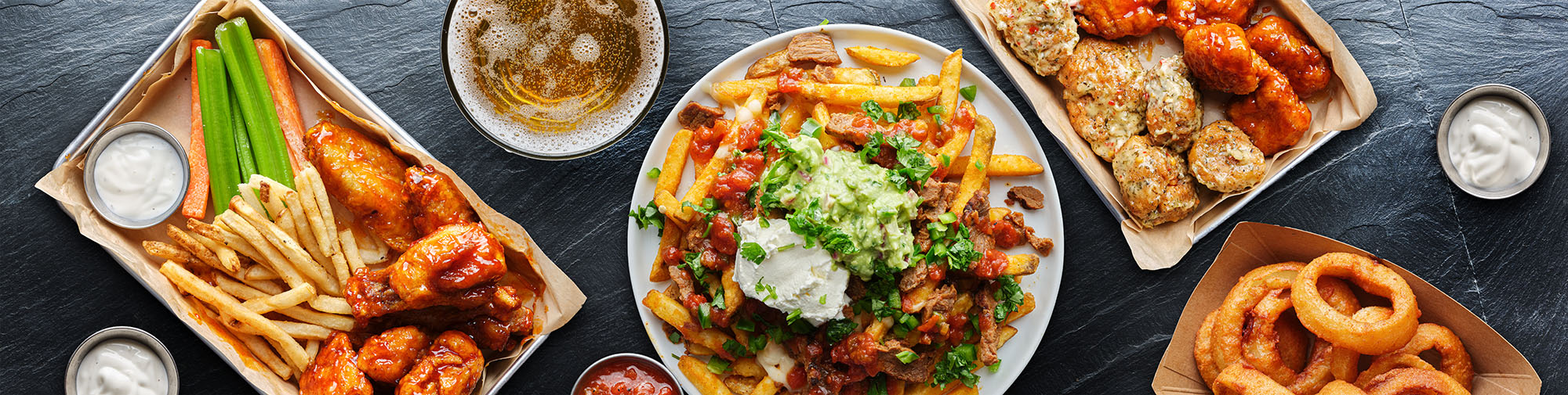 Several plates of food including French fries, wings, nachos and sauces.  