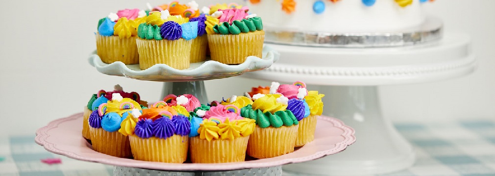 Colorful cupcakes on a tiered platter in the foreground with colorful cake in the background. 