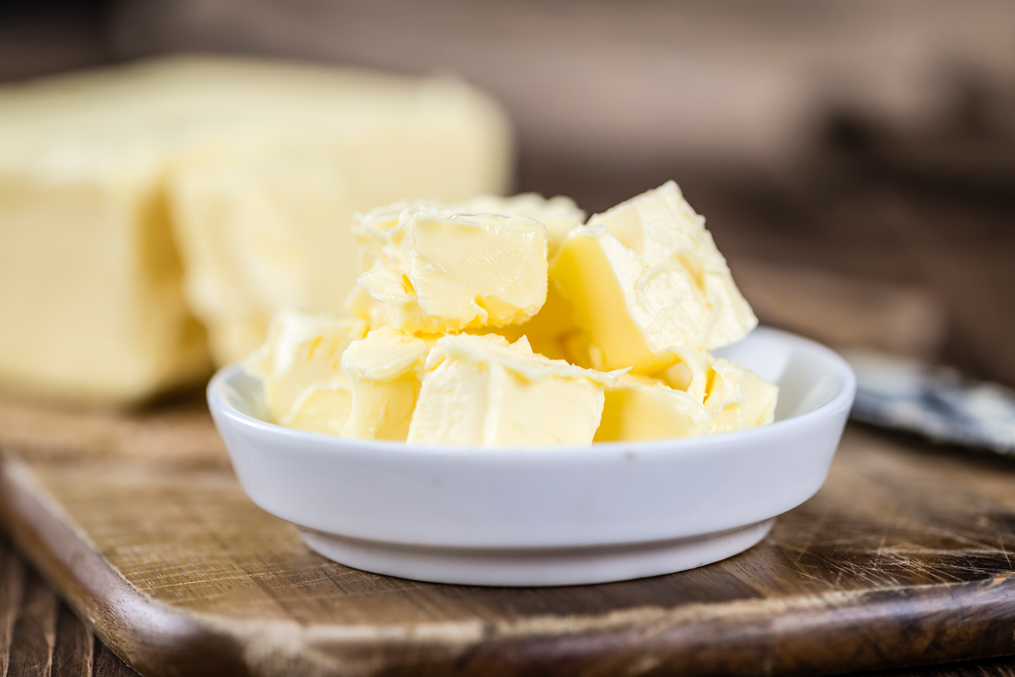 Shallow white dish with butter squares sitting in the foreground with a soft-focus block of butter behind it.  