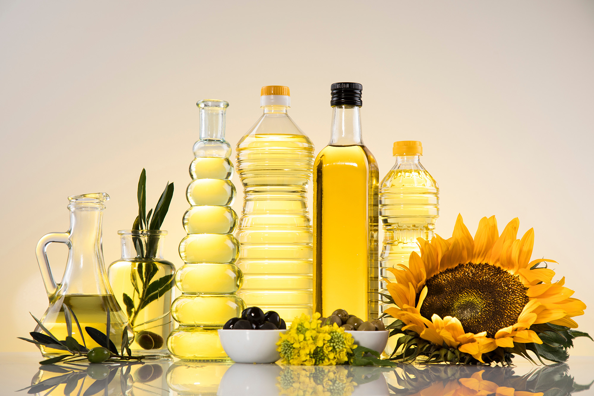Four bottles of golden oils surrounded by a sunflower, and other greens in a softly lit setting.