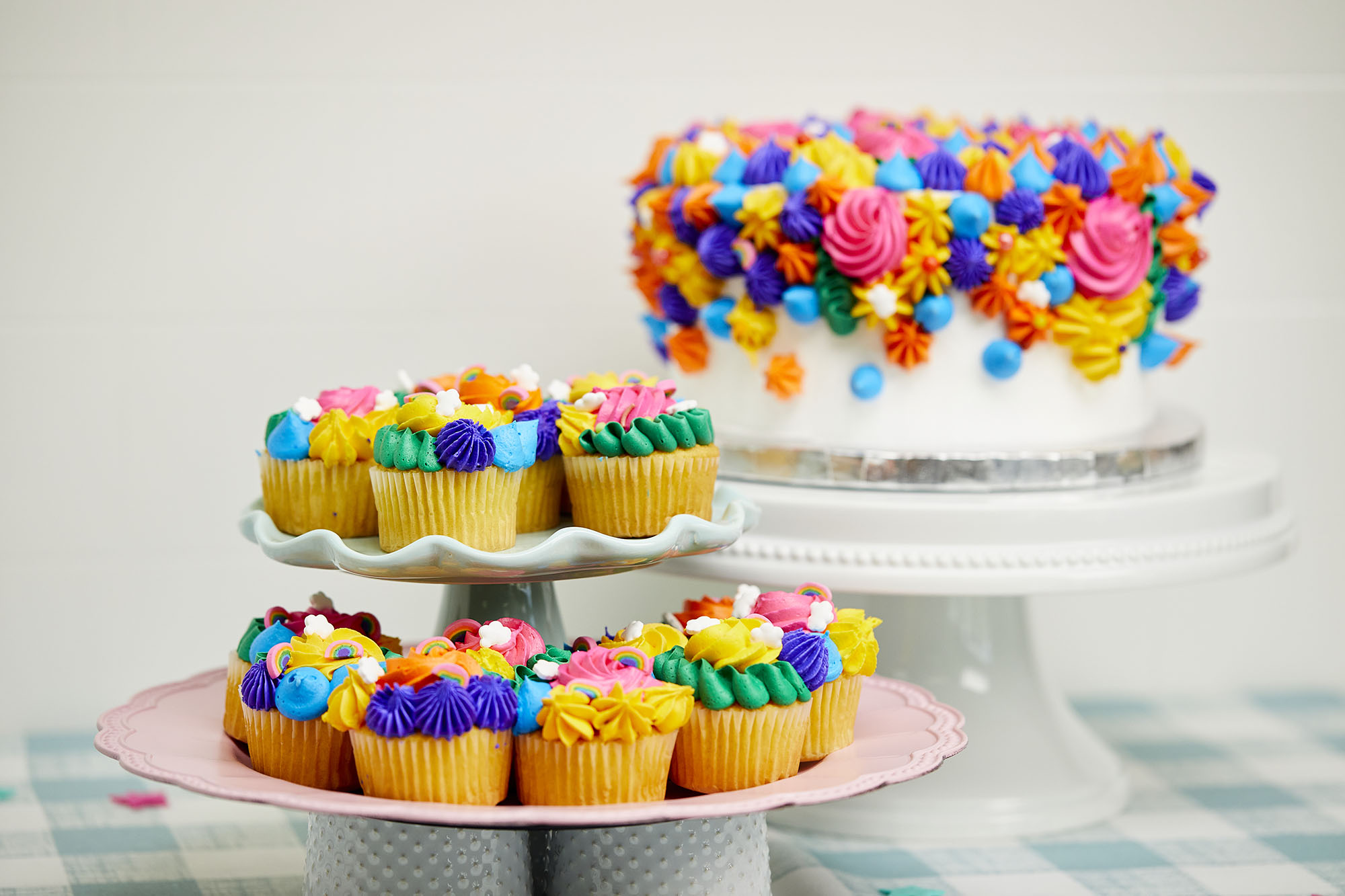Colorful cupcakes on a tiered platter in the foreground with colorful cake in the background. 
