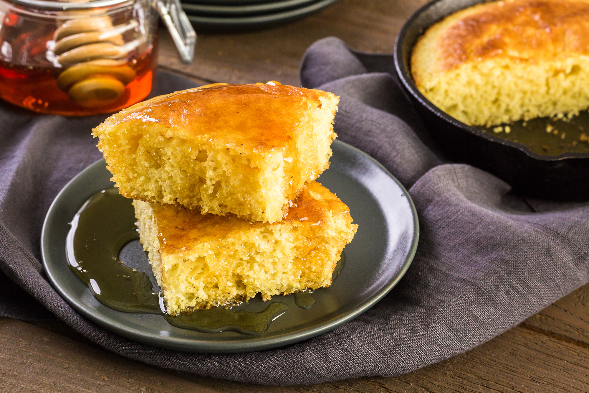 Cornbread, stacked two pieces high, rest on a dark plate. Honey drizzled over the bread.