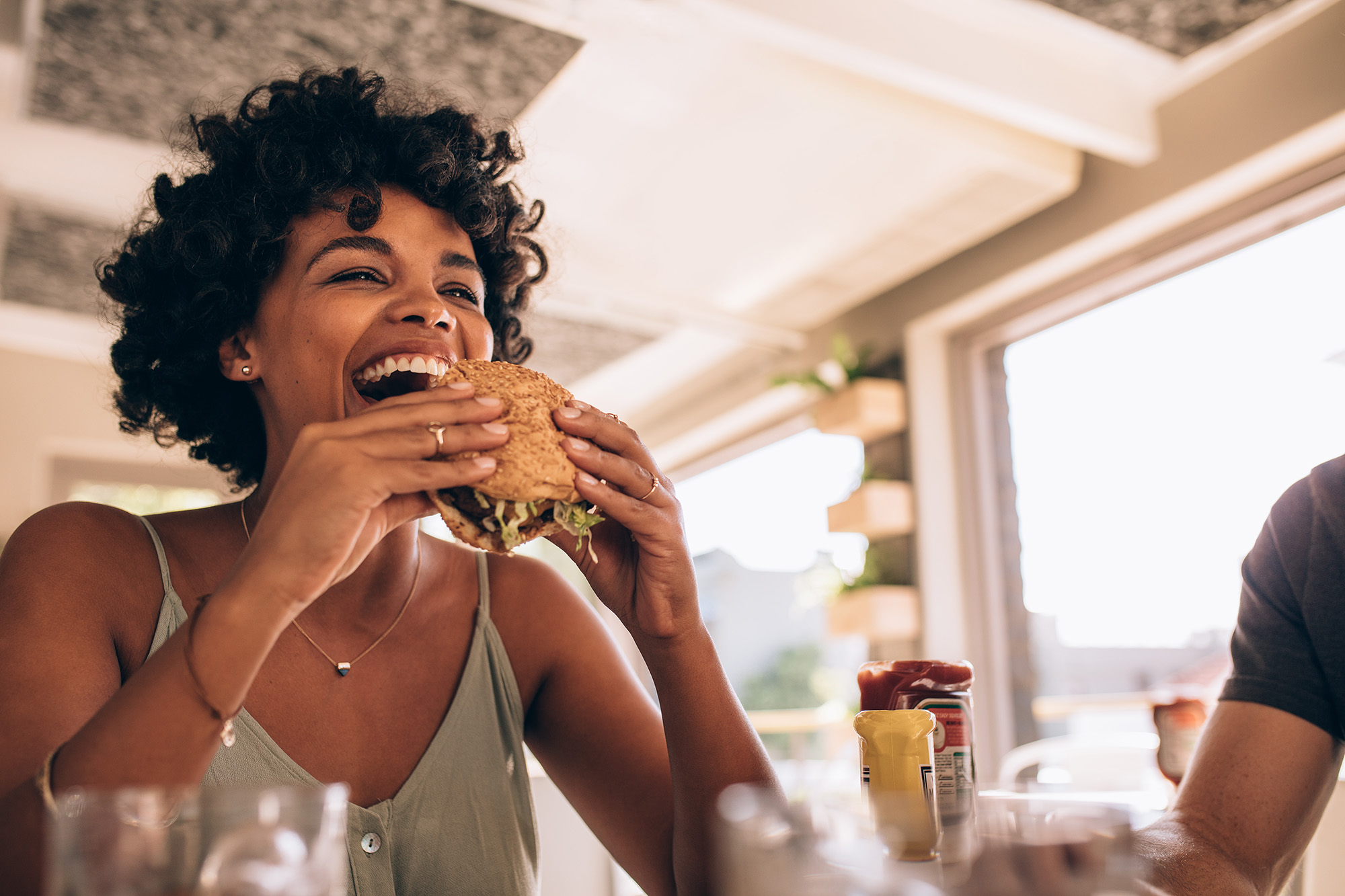 Woman sitting in a restaurant, elbows on table with her hands up to mouth. She holds a burger and is ready to dig in.
