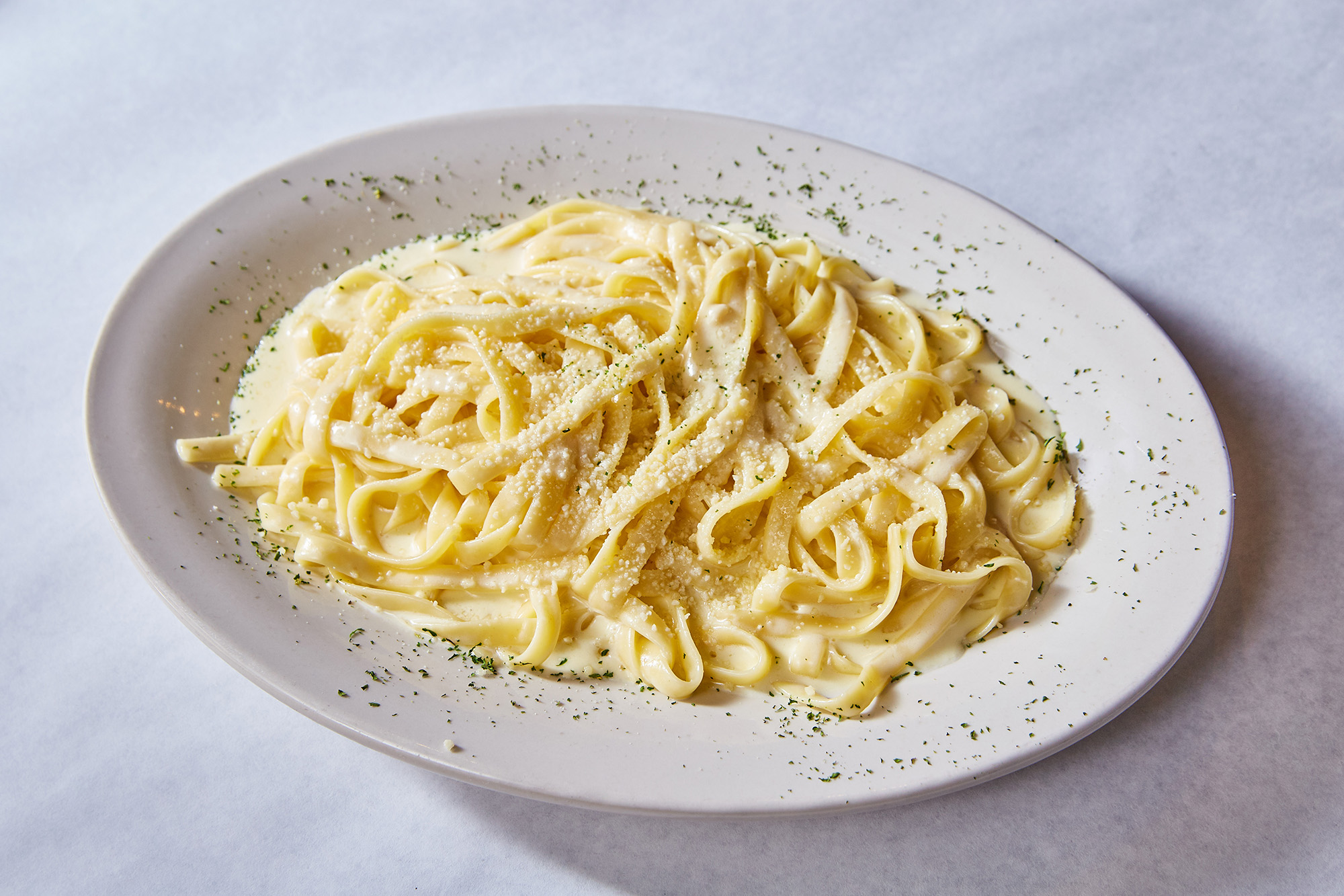A white plate of pasta alfredo with a sprinkling of garnish on top of it.