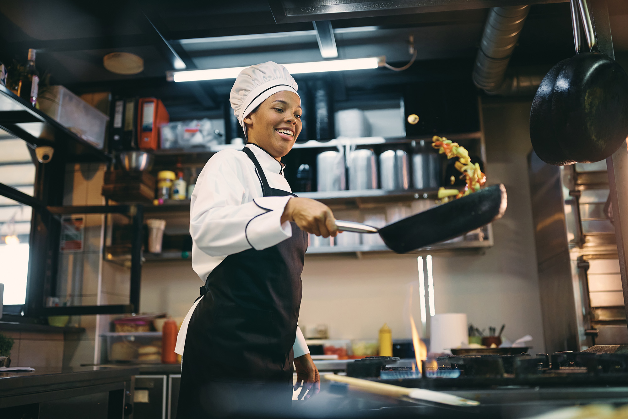 A smiling female chef in a restaurant kitchen sauteing ingredients as she flips them in the air. 